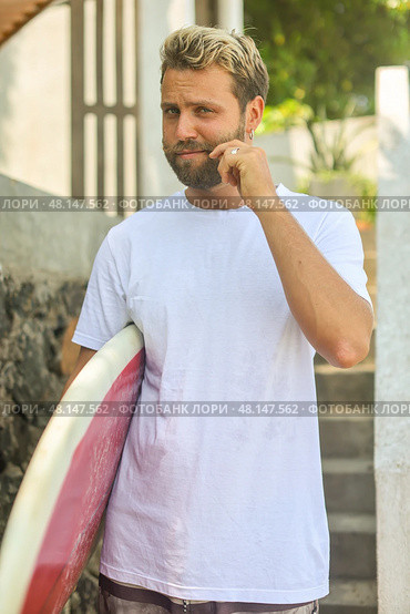 A Young Man Embracing a Surfing Lifestyle with His Surfboard in a Vibrant Tropical Environment. Стоковое фото, фотограф Женя Канашкин / Фотобанк Лори