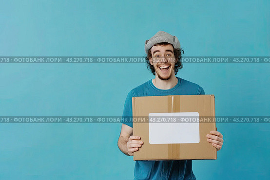 A young man is holding a cardboard box with a white label on a light blue background. The concept of delivery. Copy space. Стоковое AI-изображение, AI-художник Ярославский Максим / Фотобанк Лори