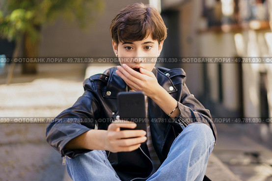 A young person displaying a shocked expression intently checks their smartphone while seated on outdoor steps in a bustling urban environment, illustrating the dynamics of modern digital engagement. A surprised young individual engaging deeply with their smartphone while sitting on urban steps. Стоковое фото, агентство Ingram Publishing / Фотобанк Лори