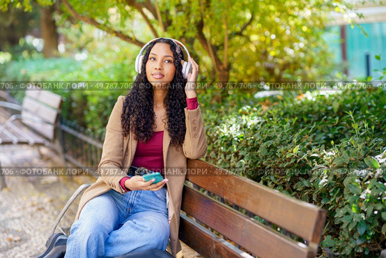 A young woman enjoys her day while sitting on a comfortable bench in a beautiful park. She is wearing stylish headphones, listening to her favorite music, and surrounded by lush, vibrant greenery. A Young Woman Delightfully Enjoying Music in a Relaxing Park Setting on a Pleasant Day. Стоковое фото, агентство Ingram Publishing / Фотобанк Лори