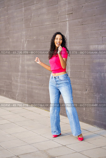 A young woman in a bright pink crop top and flared jeans dances joyfully against a textured wall, showcasing urban fashion and a vibrant lifestyle that inspires creativity and fun. A Joyful Woman is Dancing Enthusiastically in an Urban Setting, Showcasing a Trendy Outfit. Стоковое фото, агентство Ingram Publishing / Фотобанк Лори