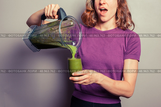 A young woman in a purple top is holding a blender with a green fruit smoothie and pouring it into a glass. Стоковое фото, агентство Ingram Publishing / Фотобанк Лори