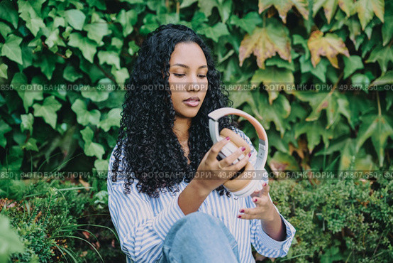 A young woman is peacefully examining and enjoying her stylish headphones, all while being surrounded by vibrant and lush greenery, which beautifully embodies a sense of tranquility and modern style. A young woman enjoys music with headphones, sitting peacefully amid vibrant green nature. Стоковое фото, агентство Ingram Publishing / Фотобанк Лори