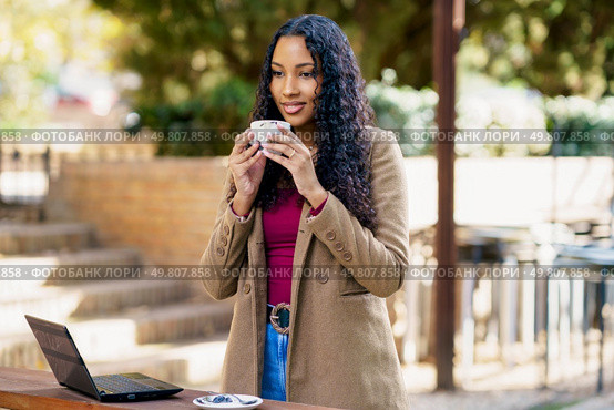 A young woman sits outside, happily sipping a warm cup of coffee as she works intently on her laptop. She embodies a perfect and relaxing worklife balance in a serene and beautiful natural setting. A young woman is savoring a delicious cup of coffee while sitting outdoors beside her laptop. Стоковое фото, агентство Ingram Publishing / Фотобанк Лори