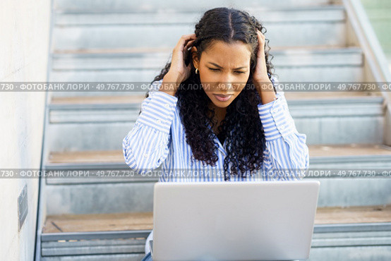 A young woman with curly hair shows frustration while using her laptop on the stairs, battling tech issues and concentration problems, leading to emotional distress and increased stress. A young woman feeling stressed and anxious as she struggles with frustrating laptop issues. Стоковое фото, агентство Ingram Publishing / Фотобанк Лори