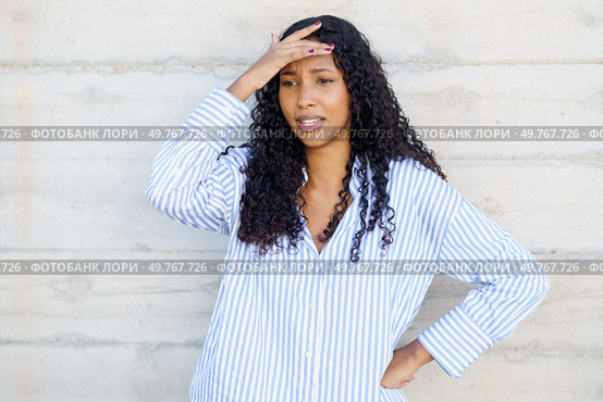 A young woman with curly hair stands pensively against a rough, concrete wall, her expression visibly stressed and deep in thought, as she wears a casual striped shirt that reflects her mood. A stressed young woman holds her head, deep in thought and emotion, facing internal battles. Стоковое фото, агентство Ingram Publishing / Фотобанк Лори