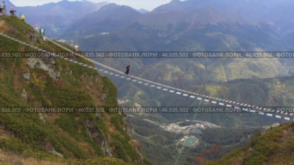 Active recreation in the Caucasus: tourists carefully walk along the suspension bridge at Rosa Peak Mountain. Активный отдых на Кавказе: Туристы осторожно идут по подвесному мосту у горы Роза Пик. Редакционное видео, видеограф Валерий Тесаков / Фотобанк Лори