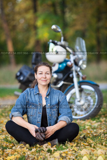 Adult biker girl sits in the lotus position in front of stylish motorcycle at autumn park, smiling happy woman on fallen leaves. Стоковое фото, фотограф Кекяляйнен Андрей / Фотобанк Лори