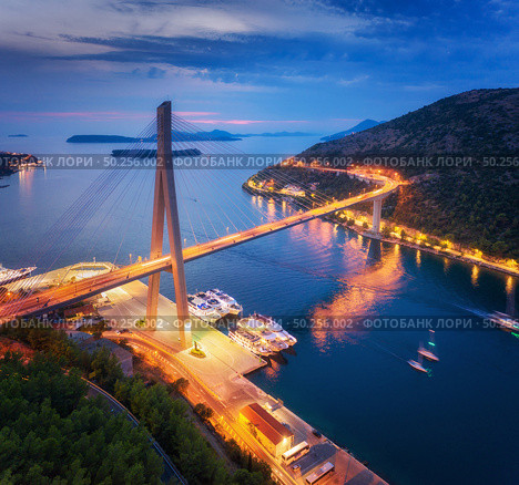 Aerial view of beautiful modern bridge at night. Dubrovnik, Croatia. Top view of road, boats, yachts and city lights at sunset. Summer landscape with harbor, mountain, highway, blue sea and cloudy sky. Стоковое фото, агентство Ingram Publishing / Фотобанк Лори