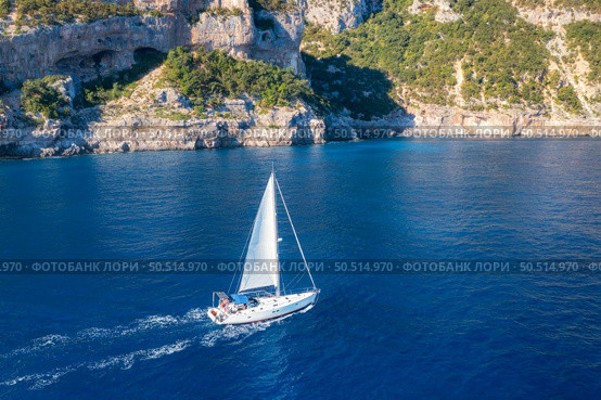 Aerial view of floating sailboat and rocky sea coast at summer sunny day. Top drone view of yacht, mountains, trees and blue water. Yachting in Sardinia, Italy. Tropical seascape with boat in ocean. Стоковое фото, агентство Ingram Publishing / Фотобанк Лори