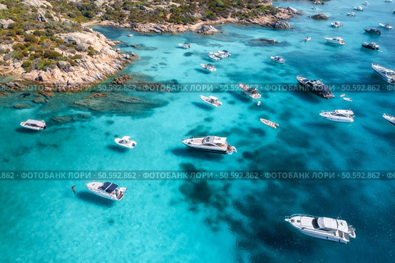 Aerial view of luxury yachts and boats navigating clear turquoise waters of the mediterranean sea near a rocky coastline with sparse vegetation. Top drone view. Summer vacation in Sardinia, Italy. Aerial view of luxury yachts and boats enjoying the crystal-clear turquoise waters of a stunning cove in sardinia, italy, creating a picturesque scene of summer leisure and idyllic escape. Стоковое фото, агентство Ingram Publishing / Фотобанк Лори