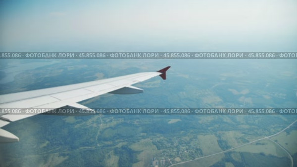 Airplane wing hovering over verdant countryside, revealing patchwork fields, rustic villages, and serpentine roads during sunny aerial journey. Стоковое видео, видеограф Mikhail Davidovich / Фотобанк Лори