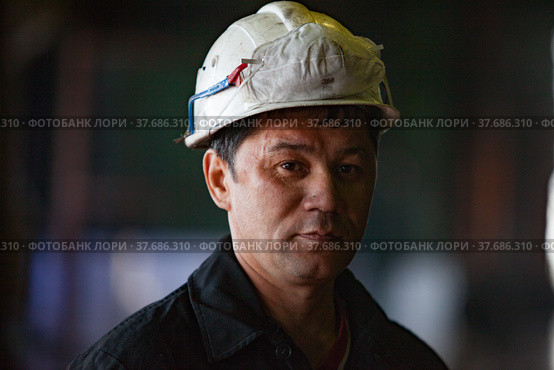 Aksu, Pavlodar region, Kazakhstan - May 29, 2012: Aksu steam power station. Portrait of adult Asian worker in white hardhat with respirator on. Close up photo. Blurred background. Редакционное фото, фотограф Alexey Rezvykh / Фотобанк Лори