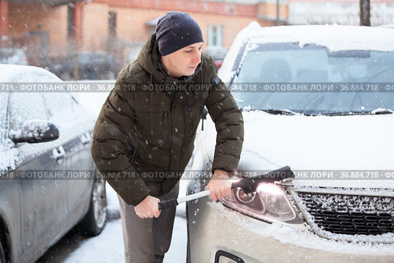 An adult Caucasian man with snow brush in hand clearing his car in winter season, swiping snow from headlight. Стоковое фото, фотограф Кекяляйнен Андрей / Фотобанк Лори