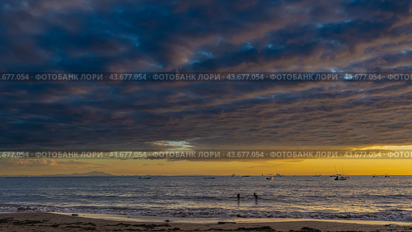 An alarming sunset over the ocean. The blue clouds are highlighted in pink. Orange sky above the horizon. Boats on the water. Silhouettes of floating people. The waves are foaming on the sandy beach. Стоковое фото, фотограф Вера Ивановна Кубарева / Фотобанк Лори