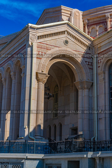An architectural fragment of the building of the cultural and historical complex "New Chersonesos" in Sevastopol. In the foreground is a massive arch with decorative carvings and columns made of light stone. The architecture contains elements of the Byzantine and neoclassical styles: arched windows, geometric ornamentation, panels and carved capitals. Clear sunlight emphasizes the relief of the details. In the background is a bright blue sky, creating a visual contrast with the beige and pink palette of the facade. The building looks monumental, emphasizing the connection with the cultural heritage of Chersonesos. Стоковое фото, фотограф Анна Кривицкая / Фотобанк Лори