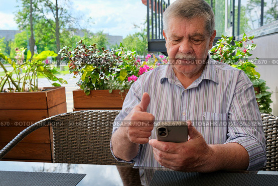 An elderly man is talking on a mobile phone near a cafe. A pensioner with a smartphone. An elderly man's emotions during a phone conversation. Стоковое фото, фотограф Терехова Марина / Фотобанк Лори