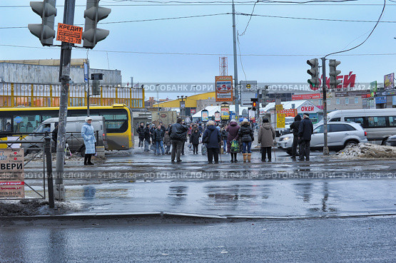 An urban scene at a busy intersection in St. Petersburg in the winter-spring period. In the foreground, wet asphalt and puddles reflecting traffic lights and pedestrians. A group of people are waiting for the green light to cross the road. Some pedestrians are wearing warm winter jackets and hats, buses, cars and shopping centers are visible in the background, including a large sign of the O'Key hypermarket. There are many billboards, store signs and street signs around. The atmosphere of everyday life in a megalopolis on a cloudy, cold day. (2013 год). Редакционное фото, фотограф Анна Кривицкая / Фотобанк Лори