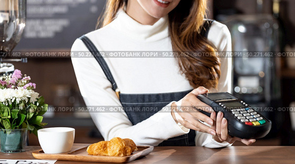 Asian Barista woman holding creadit card reader machine cashless payment for coffee purchase at cafe. Cashless preventing from coronavirus covid-19 Spreading and infection, Panoramic web banner crop. Стоковое фото, агентство Ingram Publishing / Фотобанк Лори