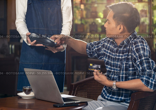 Asian customer using credit card with EMV chip technology to pay a waitress for coffee purchase at table in cafe. Стоковое фото, агентство Ingram Publishing / Фотобанк Лори
