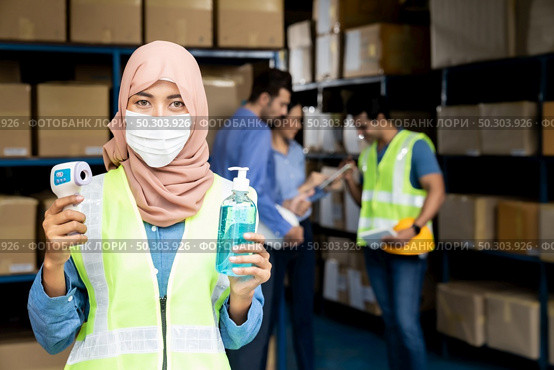 Asian Islam Muslim female warehouse worker with face mask hold hand sanitizer and infrared thermometer. For New normal hygiene life in business warehouse inventory and logistic concept. Стоковое фото, агентство Ingram Publishing / Фотобанк Лори