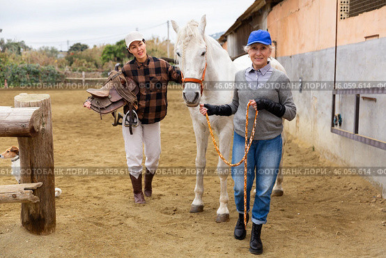 Asian woman holding saddle while older woman rancher leading horse outdoors. Стоковое фото, фотограф Яков Филимонов / Фотобанк Лори