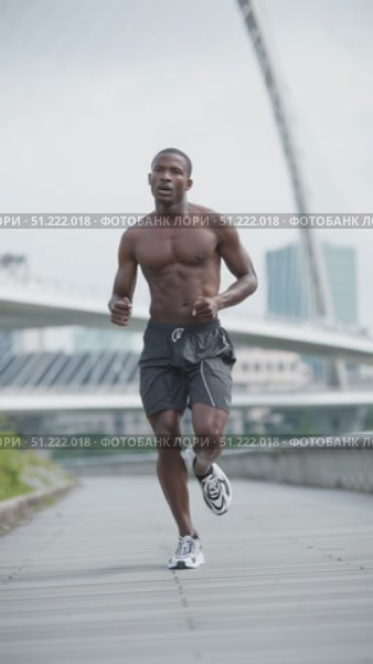Athletic black man with a muscular physique is running shirtless, wearing black shorts and sneakers, on a paved path, with a modern bridge and city skyline in the background. Стоковое AI-видео, аниматор Юрий Поздников / Фотобанк Лори
