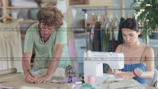 Attentive middle-aged male dressmaker drawing on paper using curves while woman sewing in workshop. Стоковое видео, видеограф Яков Филимонов / Фотобанк Лори