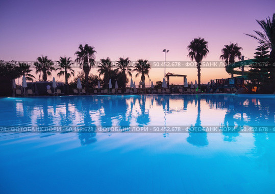 Beautiful reflection in swimming pool at colorful sunset. Purple sky reflected in water, palm trees, sun beds, umbrellas at night in summer. Luxury resort. Landscape with empty pool in twilight. Стоковое фото, агентство Ingram Publishing / Фотобанк Лори