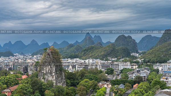 Beautiful urban and mountain scenery. City buildings surrounded by green trees. A high cliff with steep slopes and a pavilion at the top. A mountain range against the sky and clouds. China. Guilin. Стоковое фото, фотограф Вера Ивановна Кубарева / Фотобанк Лори