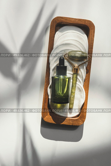 Beauty product setting with green bottle with pipette, jade massage roller and reusable cosmetic pads on white background with natural sunlight and shadow. Healthy skin care. Top view. Стоковое фото, агентство Ingram Publishing / Фотобанк Лори