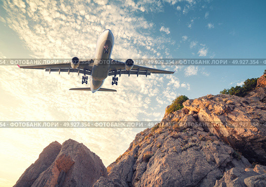 Big white aircraft is flying over rocks at sunset. Landscape with passenger airplane, mountains, colorful blue sky with clouds. Passenger airplane is landing. Business travel. Commercial plane. Стоковое фото, агентство Ingram Publishing / Фотобанк Лори