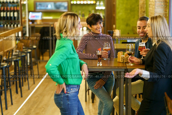 Blond female with glass of wine telling story to diverse male and female friends while gathering around table in bar together. Blond woman speaking with friends in pub. Стоковое фото, агентство Ingram Publishing / Фотобанк Лори