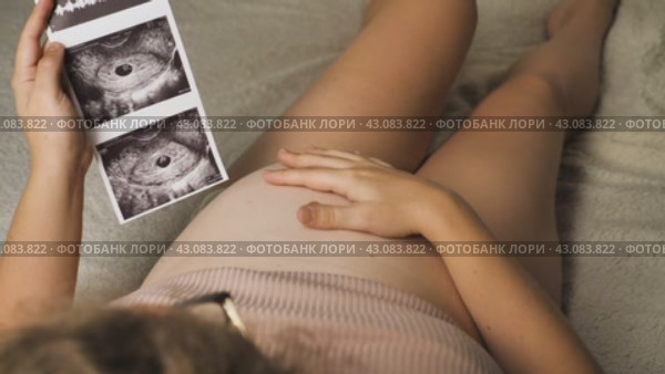 Будущая мама лежит на спине и поглаживает свой живот. Она держит в руках ультразвуковую фотографию своего ребенка - символ новой жизни, которую она вынашивает. Радость, предвкушение и эмоциональный путь материнства. Стоковое видео, видеограф Воловодов Владимир / Фотобанк Лори