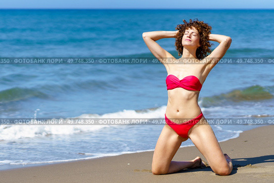 Calm slim female with curly hair in bikini standing on knees and hands behind head while getting suntan on coast. Relaxing woman sunbathing near waving sea. Стоковое фото, агентство Ingram Publishing / Фотобанк Лори