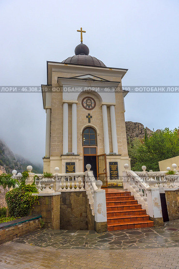 Chapel of the Nativity of Christ, located in the Laspi tract, Crimea. This is a small Orthodox chapel with classical architecture: a light facade, four columns at the entrance, a semicircular dome with a cross on top. Above the entrance is an image of Jesus Christ, and below it is a golden cross. A staircase with terracotta steps, framed by white railings with decorative balls, leads to the entrance. The open door of the chapel symbolically invites to prayer and spiritual reflection. (2021 год). Редакционное фото, фотограф Анна Кривицкая / Фотобанк Лори