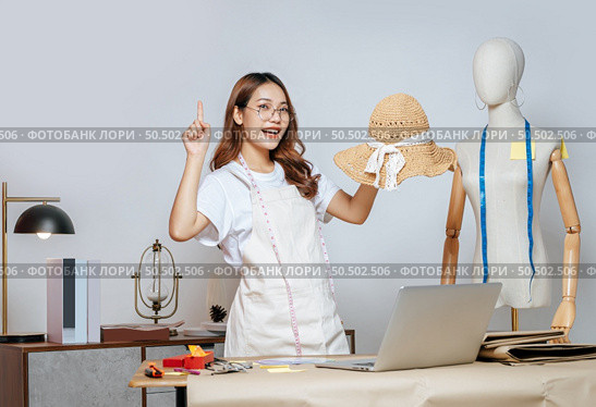 Cheerful Asian young female designer wearing eyeglasses doing fashion design on beautiful sombrero in studio, full of tailoring tools  with mannequin and equipment on the desk. Стоковое фото, агентство Ingram Publishing / Фотобанк Лори