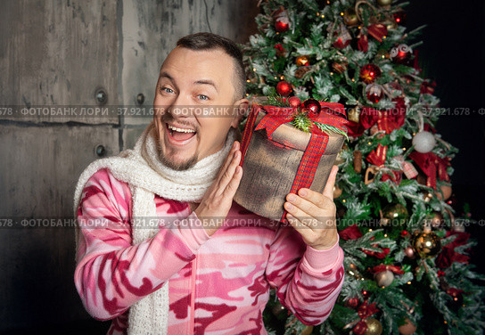 Cheerful funny happy man in pink sleepwear with Christmas present near decorated fir tree. Стоковое фото, фотограф Darkbird77 / Фотобанк Лори
