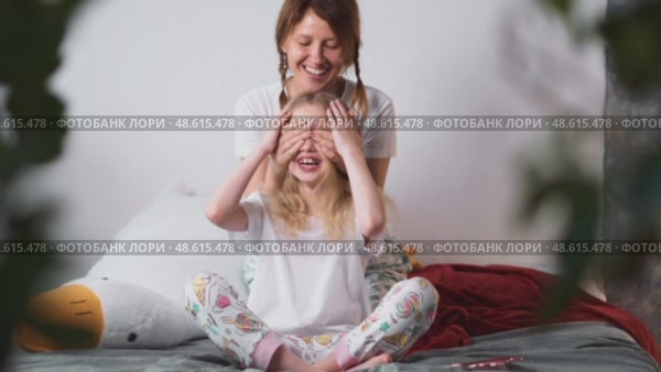 Cheerful woman gently covers her daughters eyes during play on bed. Loving moment full of emotion, care and warmth, representing pure family fun and safety. Стоковое видео, видеограф Енваев Андрей / Фотобанк Лори