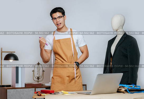 Cheerful young Asian fashion designer or tailor wearing eyeglasses use laptop computer to search idea or online orders, mannequin in suit and full of tailoring tools with equipment on desk in studio. Стоковое фото, агентство Ingram Publishing / Фотобанк Лори