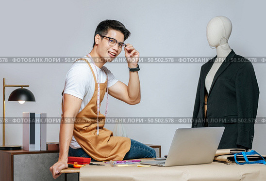 Cheerful young Asian fashion designer or tailor wearing eyeglasses use laptop computer to search idea or online orders, mannequin in suit and full of tailoring tools with equipment on desk in studio. Стоковое фото, агентство Ingram Publishing / Фотобанк Лори