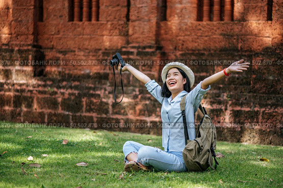 Cheerful young backpacker sitting on grass raise up her arm and smiling with happiness, holding a camera in hand while traveling in ancient site. Стоковое фото, агентство Ingram Publishing / Фотобанк Лори