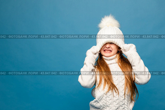 Child girl in a white sweatshirt and knitted hat with pompom on a blue background. The concept of children's emotions, joy and delight. Advertising clothing and sales. Ski resort vacation, copy space. Стоковое фото, фотограф Ирина Михайличенко / Фотобанк Лори
