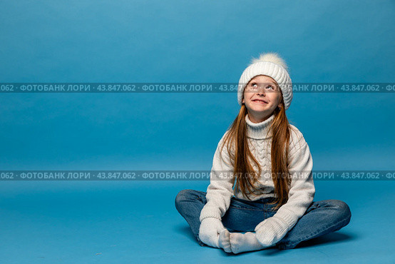 Child girl in a white sweatshirt and knitted hat with pompom on a blue background. The concept of children's emotions, joy and delight. Advertising clothing and sales. Ski resort vacation, copy space. Стоковое фото, фотограф Ирина Михайличенко / Фотобанк Лори
