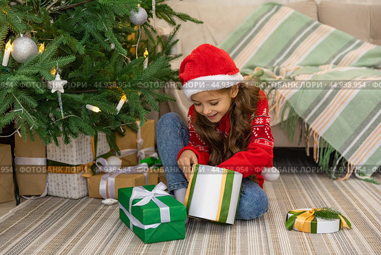 Christmas spirit. Happy little smiling girl opening gift box sitting on the floor near the Christmas tree. Happy girl unwrapping christmas present looking in box. Holidays and celebrations concept. Стоковое фото, фотограф Ирина Михайличенко / Фотобанк Лори