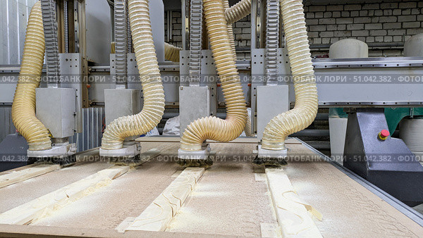 Close-up of multi-head CNC machine actively milling a workpiece, creating sawdust that is being collected by large corrugated pipes. Automated, high-precision process of modern woodworking. Стоковое фото, фотограф Елена Никишина / Фотобанк Лори