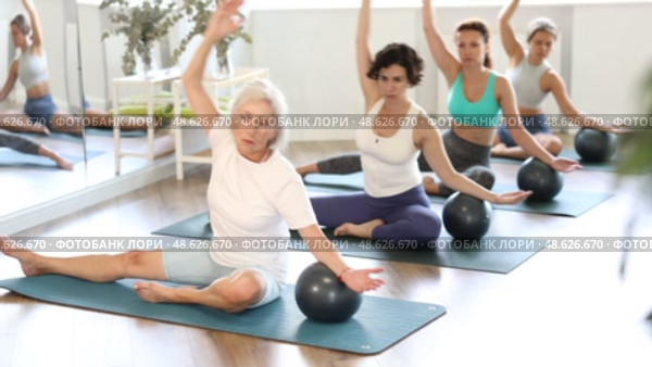 Concentrated young adult Hispanic woman, fitness enthusiast, participating group Pilates session in cozy sunlit studio, doing side bend with exercise ball, promoting balance and lateral stretching. Стоковое видео, видеограф Яков Филимонов / Фотобанк Лори