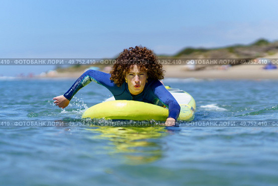 Concentrated young female surfer in wetsuit with curly hair lying on SUP board while paddling with hands floating on ocean during summer vacation. Focused woman on paddleboard rowing with hands in sea. Стоковое фото, агентство Ingram Publishing / Фотобанк Лори