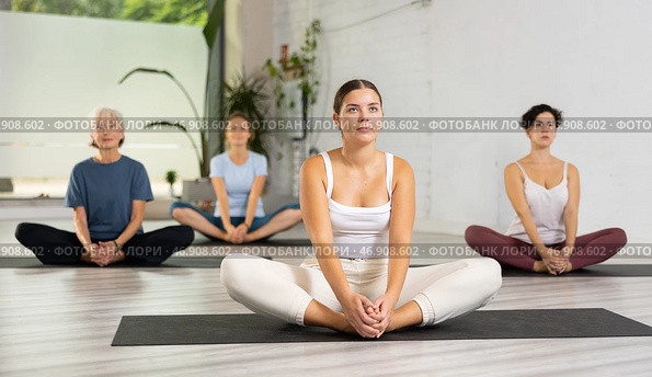 Concentrated young woman performing Butterfly Pose Baddha Konasana during yoga class with female group. Стоковое фото, фотограф Яков Филимонов / Фотобанк Лори