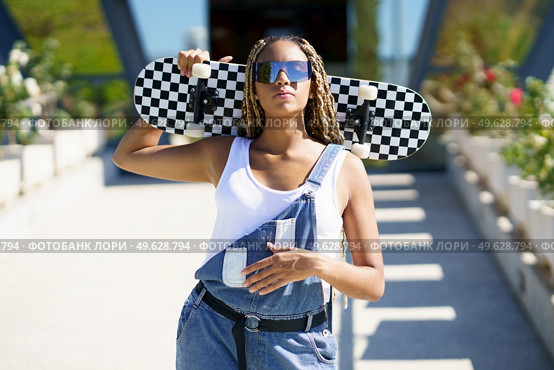 Confident African American female hipster in sunglasses and overalls standing with skateboard in street in summer and looking at camera. Cool black woman with skateboard in city. Стоковое фото, агентство Ingram Publishing / Фотобанк Лори